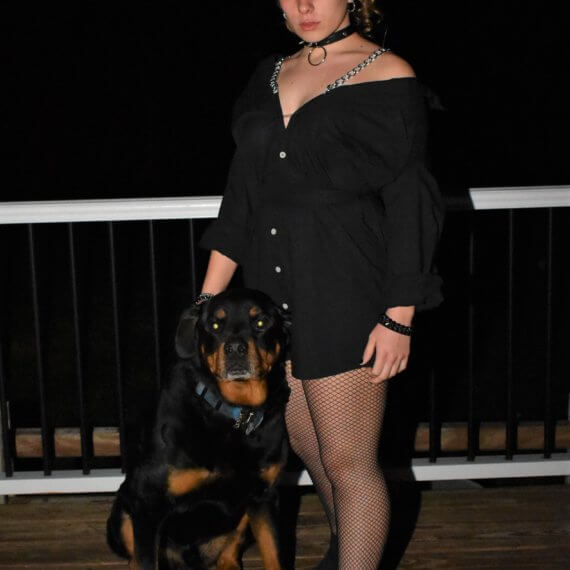 Jill Folts is modeling with rottweiler Herman while wearing a black button down shirt dress that sits off the shoulders with chain shoulder straps. This look is styled with fishnets and black boots with a spiked dog collar necklace