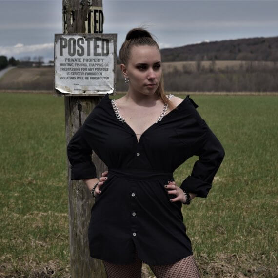 Model is wearing black shirt dress off the shoulders with chain straps. Jill is looking off to her left with her hands on her hips. She is standing in front of a “posted” sign