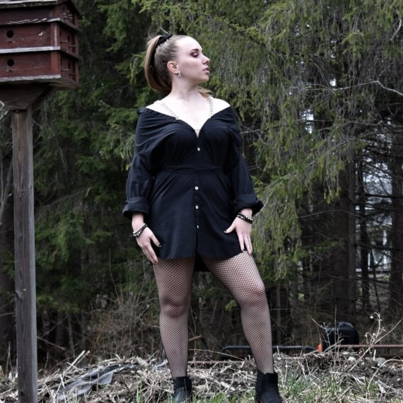 Jill is wearing a black button down shirt dress that sits off the shoulders with chain shoulder straps. This look is styled with fishnets and black boots with spiked bracelets on each wrist. She is in a wooded setting looking off to her left