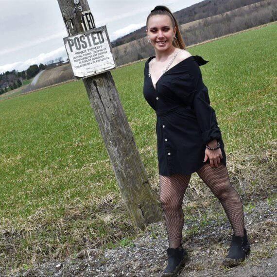 Model is wearing black shirt dress off the shoulders with chain straps. Jill is looking off to the camera smiling. She is standing in front of a “posted” sign