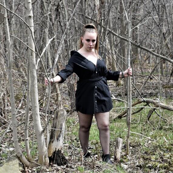 Jillian is wearing black shirt dress off the shoulders with chain straps. Jill is in the woods holding on to two small trees, one to each side of her giving a stern look to the camera