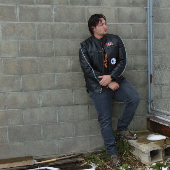 Zach Hellwig is modeling in a black leather jacket that has three patches sewn on, the Austria flag, a cartoon eye, and a red and black nautical star. There are grey chains on each sleeve. This jacket is styled with a black bleached shirt and grey pants. He has one leg on a step while leaning against a wall, looking off to his left.