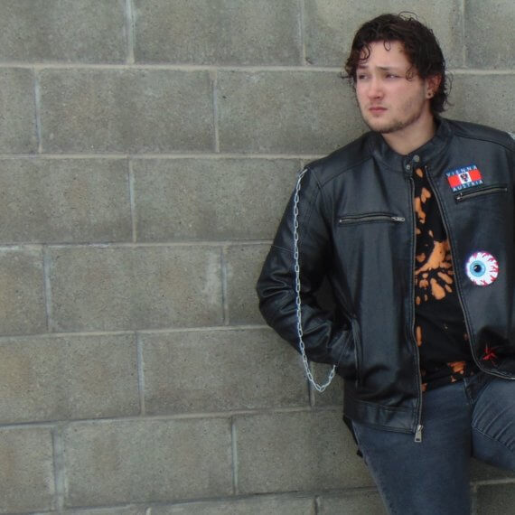 Zach is modeling in a black leather jacket that has three patches sewn on. He is looking to his right with a concerned look and both hands in his jacket pockets