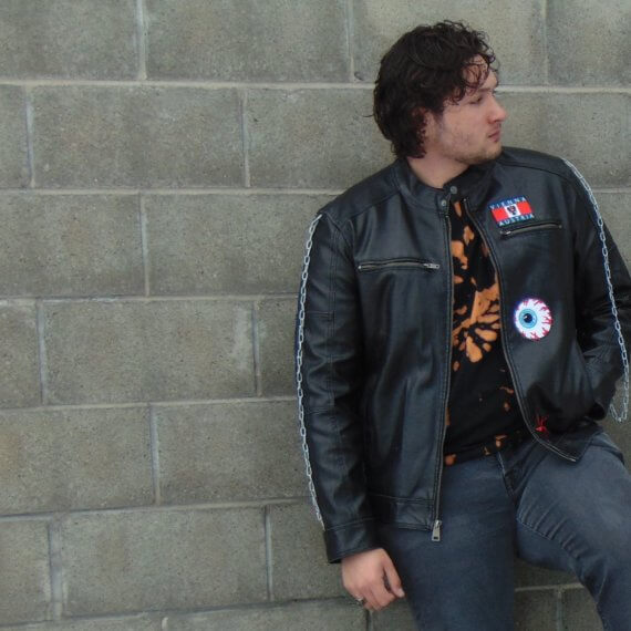 Model is in a black leather jacket that has three patches sewn on. He is looking to his left, one hand in his jacket pocket and one is down to his side
