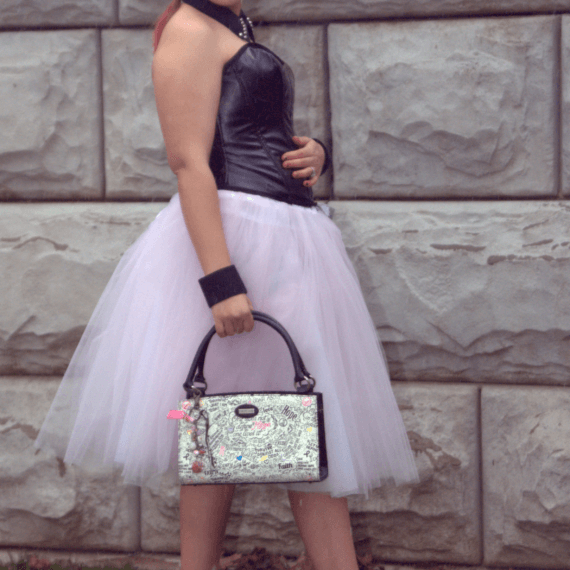 Black pleather sweetheart corset with midi length pink tulle skirt styled with black ankle boots, an upcycled black shirt collar necklace, and upcycled wrist cuffs. Alexis is turned from the back and holding onto a white purse with pink and black writing