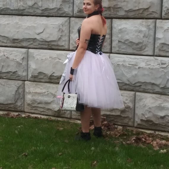 Model is turned from the back showing the lacing of the black pleather corset and of the pink tulle skirt. She is holding a white and black purse with charms and pink and black writing on it