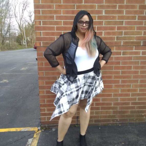 Model Shana Williams is modeling an open black sheer hooded top with a white tank top underneath with a black and white plaid handkerchief skirt styled with black ankle boots. She has her hands on her hips and the skirt is blowing in the wind