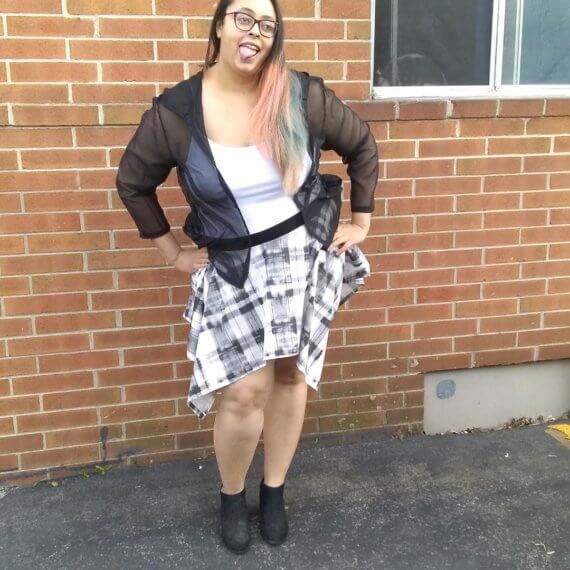 Shana is wearing a black sheer hooded top with a white tank top underneath with a black and white plaid handkerchief skirt. She has her hands on her hips and is sticking her tongue out making a funny face