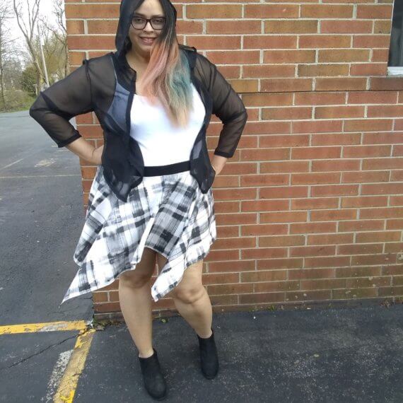 Model Shana Williams is modeling an open black sheer hooded top with a white tank top underneath with a black and white plaid handkerchief skirt styled with black ankle boots. She is smiling with her hands on her hips and the skirt is blowing in the wind