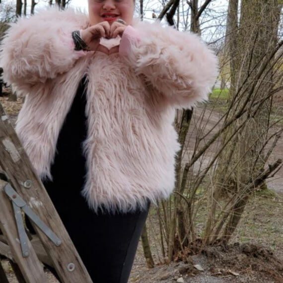 Aaries Fitzsimmons modeling a plush pink bomber jacket styled with black leggings and a black shirt. She is peeking out from behind a tree giving heart hands
