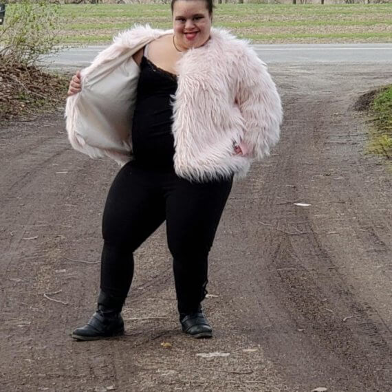 Model Aaries wearing a plush pink bomber jacket styled with black leggings and a black shirt and black ankle boots. Aaries is holding the right side of the jacket open