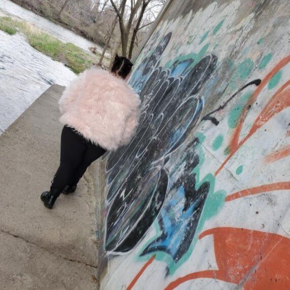 Aaries walking away from the camera showing the back of the plush pink jacket. She is walking next to a graffitied wall