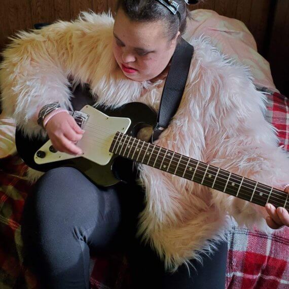 Model sitting on bed playing guitar while wearing a pink fur jacket styled with a pleather headband with spikes
