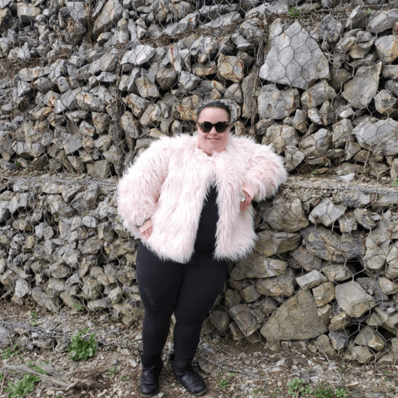 Aaries standing in front of a cobblestone wall modeling a pink fur coat, styled with black leggings, a big gem ring, sunglasses, and a pleather spike headband