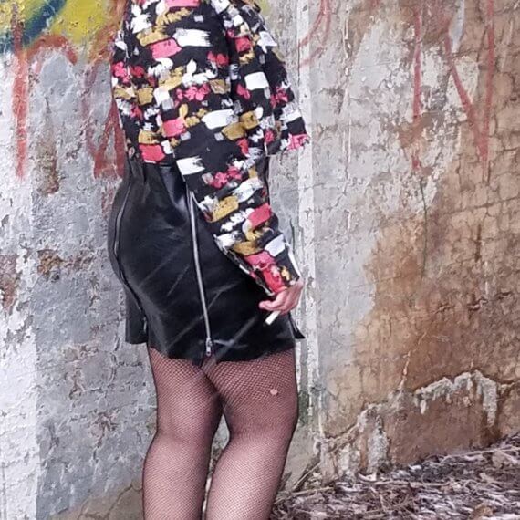 Alexis is looking over her right shoulder wearing a black distressed cropped denim jacket that is painted with a black pleather mini skirt with zippers