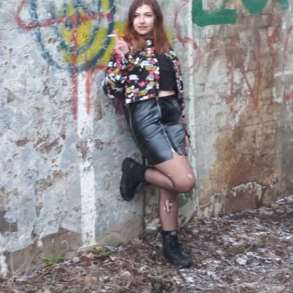 Modeling wearing a black distressed cropped denim jacket that is painted with a black pleather mini skirt with zippers. This is styled with ripped fishnets and black combat boots. Alexis is holding a fake cigarette while leaning against a wall and looking away from the camera