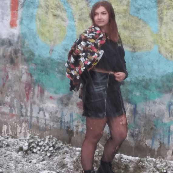 Alexis is looking over her shoulder wearing a black distressed cropped denim jacket that is painted with a black pleather mini skirt with zippers. She has the jacket half off