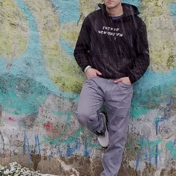 Model Ryan Markwith is leaning against a graffiti wall wearing a black hoodie with “If you’re not angry, you’re not paying attention” written on it. This is styled with grey jeans and white Doc Martin boots