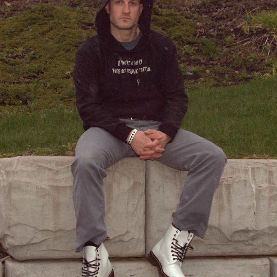 Ryan is wearing a black hoodie with the hood up styled with grey pants and white boots. He’s sitting on a stone ledge