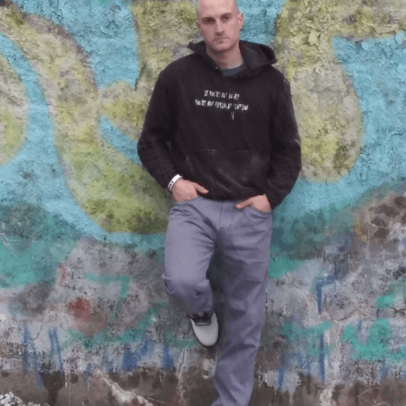 Model Ryanis leaning against a graffiti wall with his hands in his pockets looking away from the camera wearing a black hoodie. This is styled with grey jeans and white Doc Martin boots