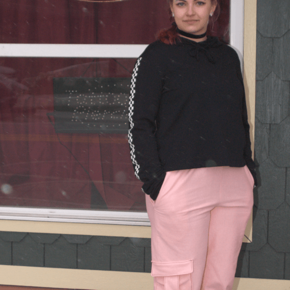 Pink sweatpants styled with a black hoodie with checkerboard stripes down the sleeve and a black velvet choker necklace. Model Alexis Remington is standing in front of Revelation Ink Tattoos and Piercings with her hands in her pockets