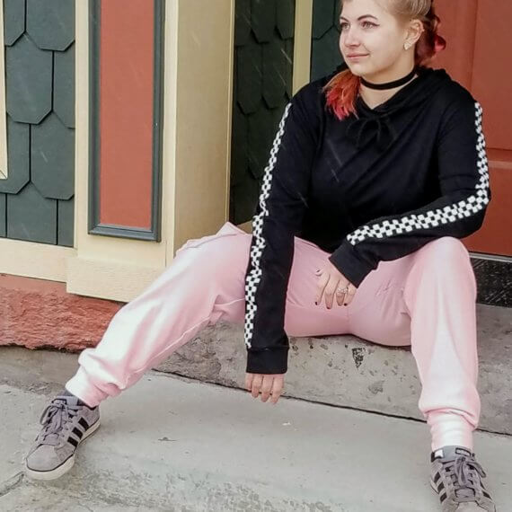 Alexis is sitting on a step looking away from the camera in pink sweatpants styled with a black hoodie with checkerboard stripes down the sleeve and a black velvet choker necklace and grey Adidas sneakers