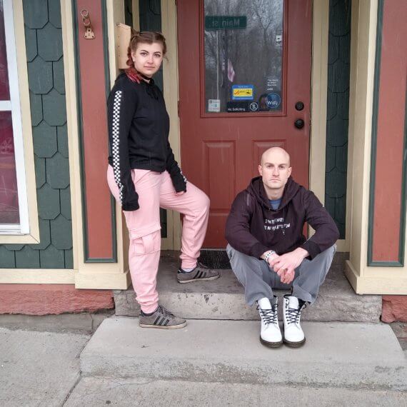 Model Alexis wearing pink sweatpants styled with a black hoodie with checkerboard stripes down the sleeve. She is leaning against a wall with one leg up on a step. Model Ryan Markwith is sitting on the step next to her.