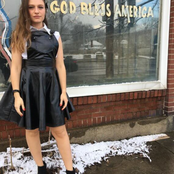Model Lindsy VonOhlen is wearing a black pleather overall dress with a sleeveless white button up shirt with spikes on the collar. This is styled with black ankle boots and a black spiked bracelet. She is standing in front of a window with “God bless America” painted on it
