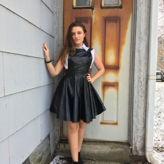 Lindsy stands in front of a rusted door with her hand on a wall modeling a black pleather overall dress with a white tank top button up shirt