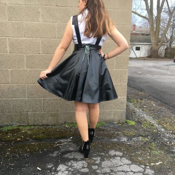 Model is standing showing off the back of the pleater overall dress. She is holding the side of the left side of her dress up. Lindsy is wearing a pleather bow hairpiece