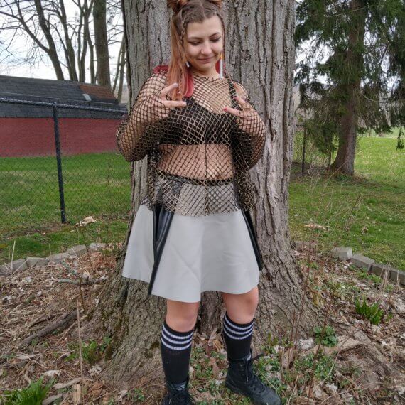 Model Alexis Remington is wearing a gold long sleeve fishnet top paired with a grey leather skirt with two black leather stripes down each side. Styled with knee high black socks and army combat boots and fake cigarette earrings. She is looking down and giving “peace signs”