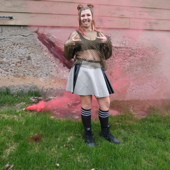 Alexis in a gold long sleeve fishnet oversized top paired with a grey leather skirt with two black leather stripes down each side. Styled with knee high black socks and army combat boots and fake cigarette earrings. Model Alexis Remington is using a red smoke bomb while giving “peace signs” and sticking out her tongue