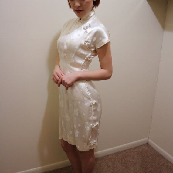 Red hair woman wearing white silk satin mid thigh length short sleeve dress with white floral embroidered overlay. The buckle slanted down from the neck to the bottom of the skirt.