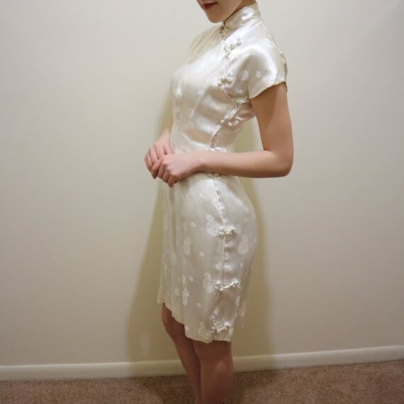 Red hair woman wearing white silk satin mid thigh length short sleeve dress with white floral embroidered overlay. The buckle slanted down from the neck to the bottom of the skirt.