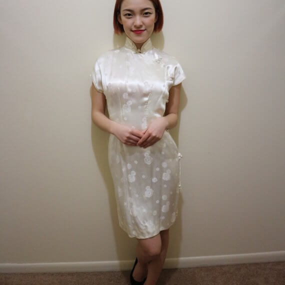 Red hair woman wearing white silk satin mid thigh length short sleeve dress with white floral embroidered overlay. The buckle slanted down from the neck to the bottom of the skirt.