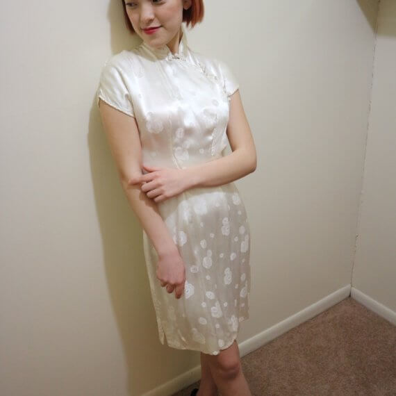 Red hair woman wearing white silk satin mid thigh length short sleeve dress with white floral embroidered overlay. The buckle slanted down from the neck to the bottom of the skirt.