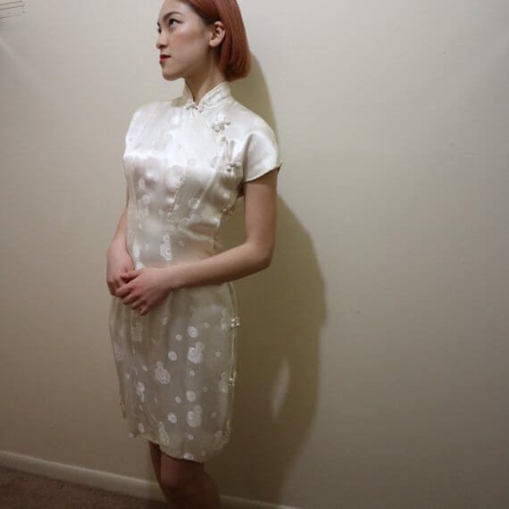 Red hair woman wearing white silk satin mid thigh length short sleeve dress with white floral embroidered overlay. The buckle slanted down from the neck to the bottom of the skirt.