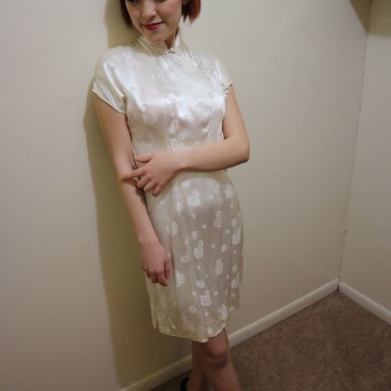 Red hair woman wearing white silk satin mid thigh length short sleeve dress with white floral embroidered overlay. The buckle slanted down from the neck to the bottom of the skirt.