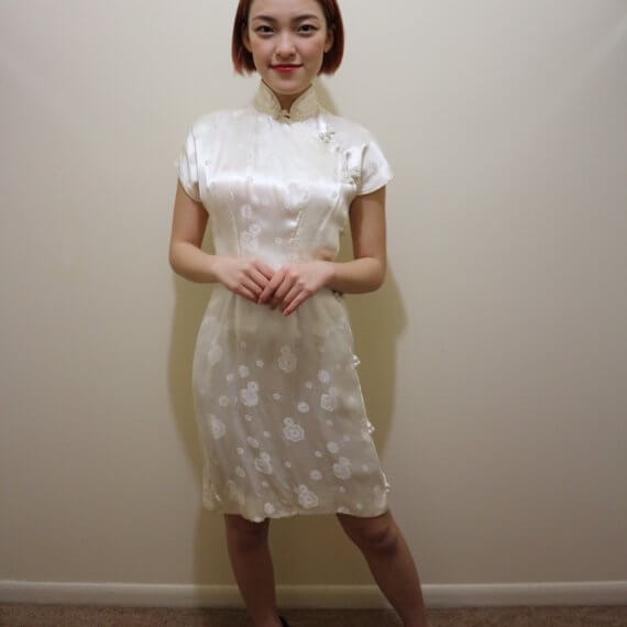 Red hair woman wearing white silk satin mid thigh length short sleeve dress with white floral embroidered overlay. The buckle slanted down from the neck to the bottom of the skirt.