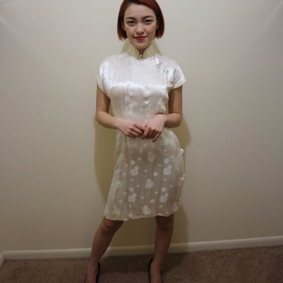 Red hair woman wearing white silk satin mid thigh length short sleeve dress with white floral embroidered overlay. The buckle slanted down from the neck to the bottom of the skirt.