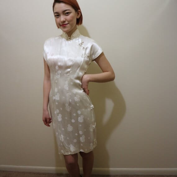 Red hair woman wearing white silk satin mid thigh length short sleeve dress with white floral embroidered overlay. The buckle slanted down from the neck to the bottom of the skirt.