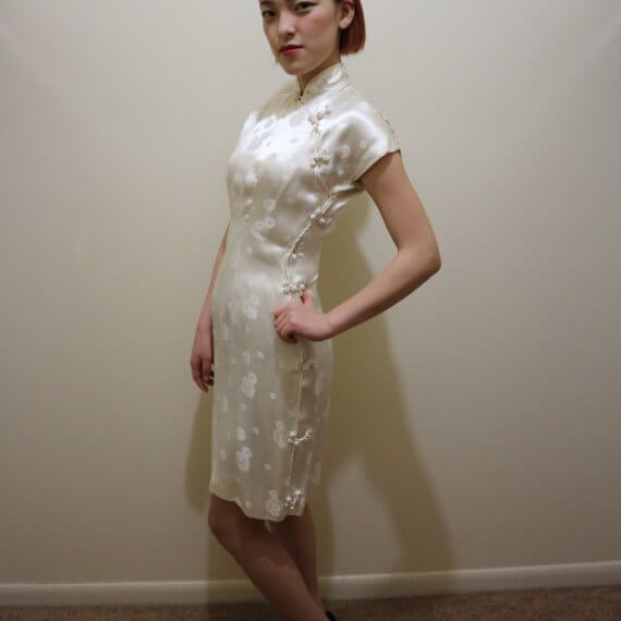 Red hair woman wearing white silk satin mid thigh length short sleeve dress with white floral embroidered overlay. The buckle slanted down from the neck to the bottom of the skirt.