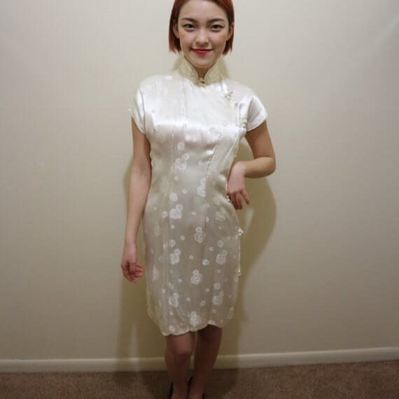 Red hair woman wearing white silk satin mid thigh length short sleeve dress with white floral embroidered overlay. The buckle slanted down from the neck to the bottom of the skirt.