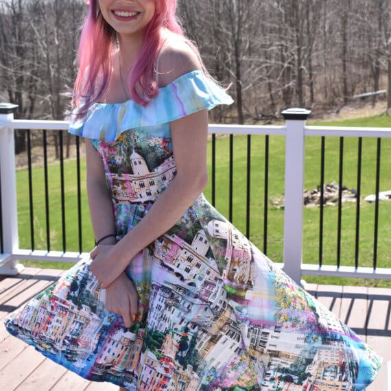 An off the shoulder, high-low sundress with pockets being blown by the wind. The fabric is printed with a clear blue canal, multicolored buildings, and a light blue sky to create a scene of Venice, Italy. The model is posing in front of a woodsy and grass filled background.