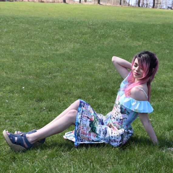 An off the shoulder, high-low sundress with pockets, paired with strappy denim wedges. The fabric is printed with a clear blue canal, multicolored buildings, and a light blue sky to create a scene of Venice, Italy. The model is sitting on a grassy hill, leaning on one arm with the other hand in her hair.