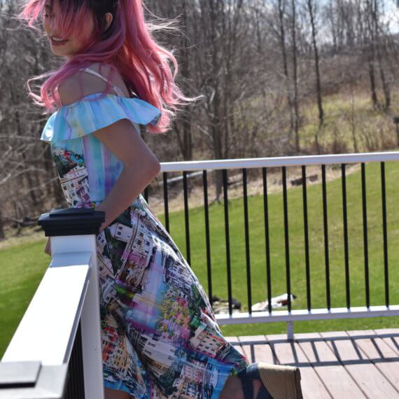An off the shoulder, high-low sundress with pockets, paired with strappy denim wedges. The fabric is printed with a clear blue canal, multicolored buildings, and a light blue sky to create a scene of Venice, Italy. The model is leaning on a porch railing with one leg kicked up behind her and her black to pink ombre hair blowing in the wind.