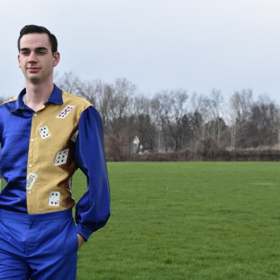 A men’s slim-fit dress shirt with a straight point collar and pirate sleeves, paired with royal blue dress pants. The dress shirt has a color blocked design to it with the right shirt front, left sleeve, sleeve cuffs, collar, and back in royal blue; left shirt front and yoke in gold, with cascading playing cards on the left shirt front; the right sleeve is white with hand written music from “La regata veneziana” by Gioacchino Rossini. The model is standing with his hands in his pockets against a woodsy and grass filled background