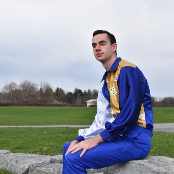 A men’s slim-fit dress shirt with a straight point collar and pirate sleeves, paired with royal blue dress pants. The dress shirt has a color blocked design to it with the right shirt front, left sleeve, sleeve cuffs, collar, and back in royal blue; left shirt front and yoke in gold, with cascading playing cards on the left shirt front; the right sleeve is white with hand written music from “La regata veneziana” by Gioacchino Rossini. The model is sitting on a rock wall in front of a grassy background