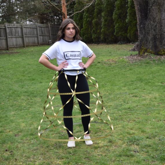 An oversized white t-shirt with black accents on the collar and cuffs, and “Casual” printed across the front, paired with black leggings and white tennis shoes. The outfit is then accessorized with a golden metal and plastic cage hoop skirt designed with horizontal and criss-cross lines. The model is standing with her hands on her hips in front of a natural background