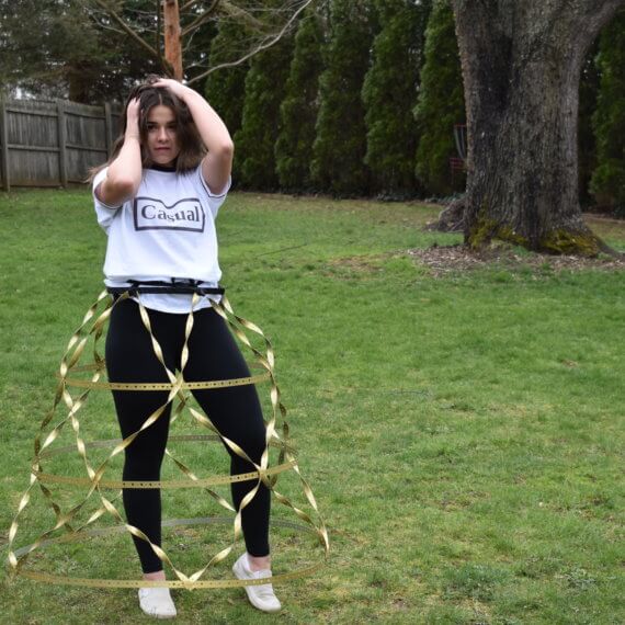 An oversized white t-shirt with black accents on the collar and cuffs, and “Casual” printed across the front, paired with black leggings and white tennis shoes. The outfit is then accessorized with a golden metal and plastic cage hoop skirt designed with horizontal and criss-cross lines. The model is posed with her hands in her hair against a natural background.